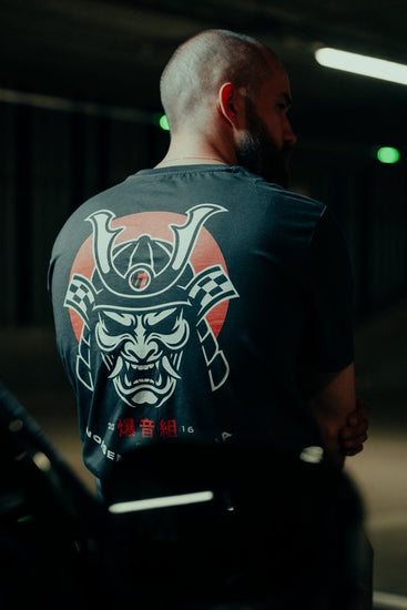 Man wearing a grey t-shirt with a samurai design on the back in a dark setting.