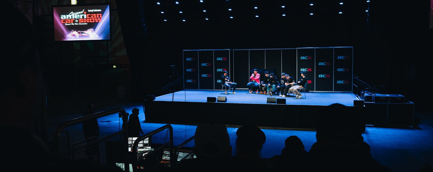 Stage with performers and audience in a darkened auditorium, featuring a screen displaying event details.